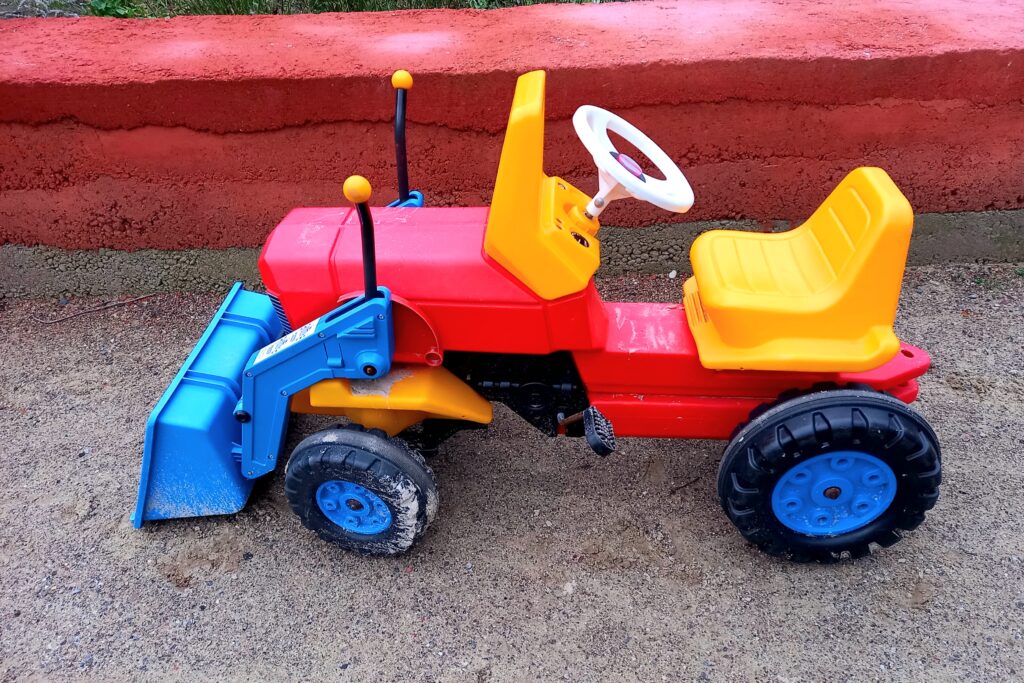 ein bunter Kinderbagger, der Bagger selbst ist rot, der Sitz gelb und die Schaufel blau; er steht auf einem Sandweg vor einer niedrigen Mauer aus Tonstein